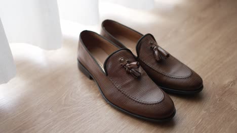 pair of brown tassel loafers placed neatly on a wooden floor by a sheer curtain