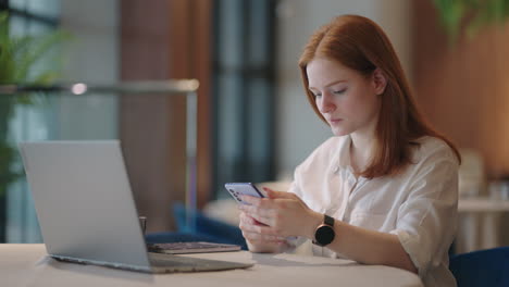 una mujer independiente está trabajando con una computadora portátil en un café enviando mensajes por teléfono inteligente y sonriendo