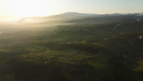 aerial drone view of the icelandic highlands at sunrise, showcasing mist-covered green fields, distant mountains, and soft sunlight over the landscape