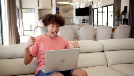 a young biracial man uses laptop on a beige sofa, celebrating a sports victory