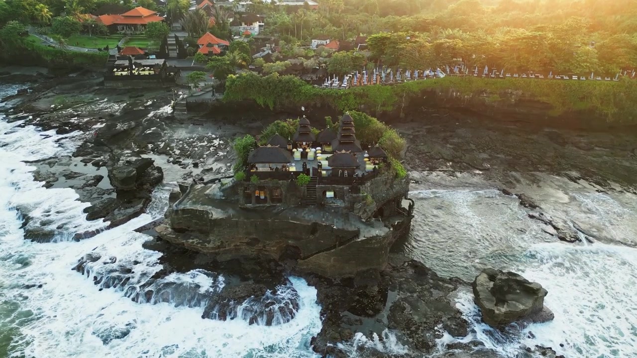 Chrám Tanah Lot tyčící se na dramatickém skalnatém pobřeží obklopený tříštícími se oceánskými vlnami a zlatým světlem východu slunce s tropickou balijskou scenérií, nájezdové letecké záběry z dronu.