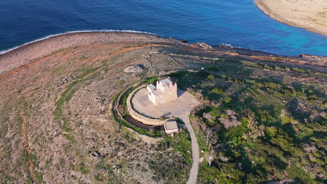 Vista aerea orbitale intorno alla restaurata Torre Bianca a Mellieha, Malta, che mette in risalto la sua architettura medievale, i paesaggi costieri e le acque blu vibranti del Mar Mediterraneo.