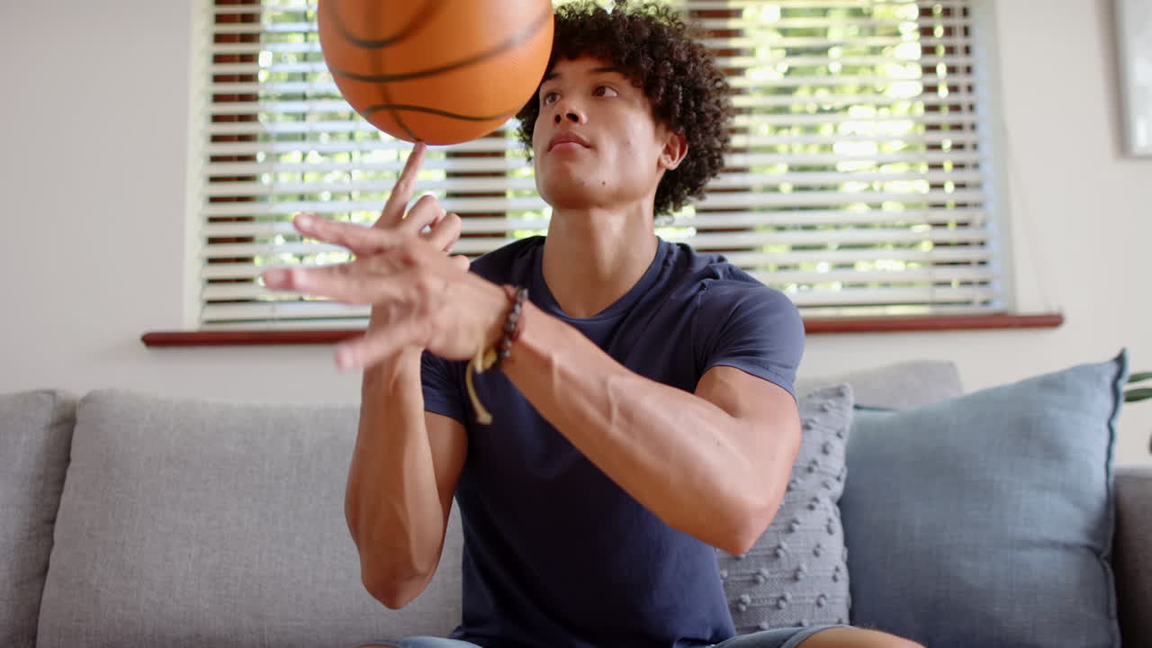 Spinning basketball on finger, young man sitting on couch, enjoying leisure time, at home