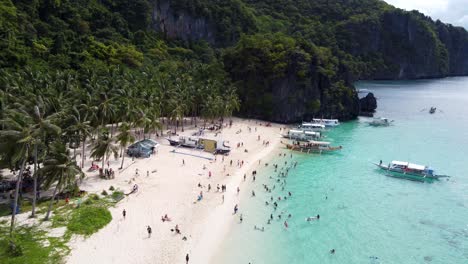 aerial: filipino outrigger tour boats anchored on tropical seven commandos beach, tourists swimming in turquoise clear water on white sand, palm trees and lush green limestone mountains