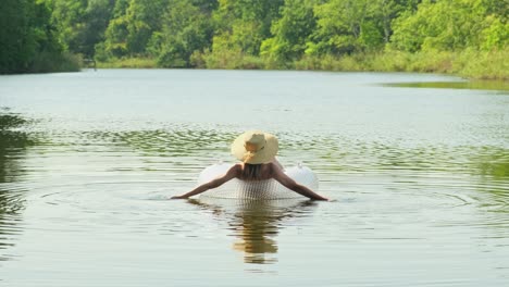 woman in swimsuit and straw hat is floating with an inflatable circle in lake river pond. girl rests on swimming ring in a pool with blue water. concept of travel, vacation, weekends, holidays, relaxing on nature, rest, relaxation