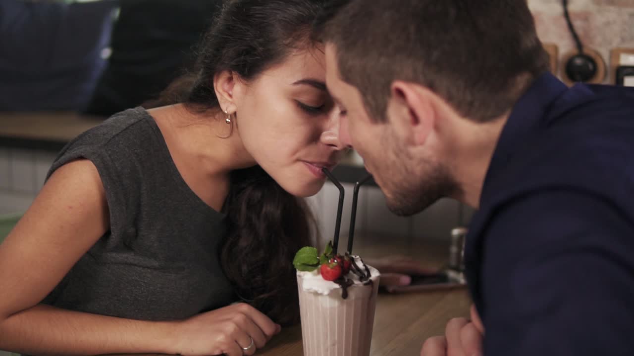 Close Up view of attractive young couple in cafe sitting at the wooden table and sharing milkshake drinking it together using two