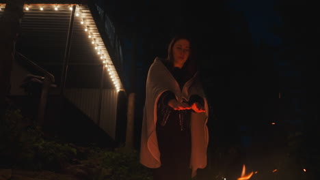 woman rolls hands over fire near forest. lady draped in snug blanket endeavors to stave off evening chill as cold night unfolds. woman enveloped in darkness