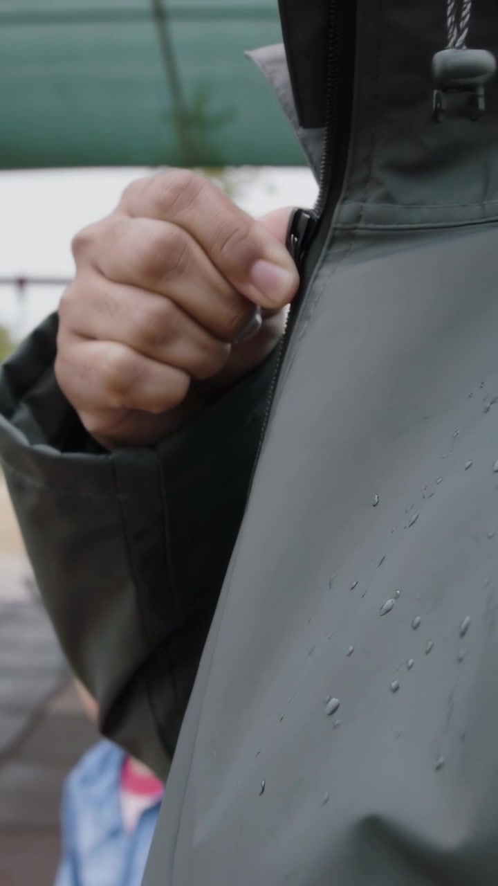 Video vertical: Hombre subiendo la cremallera de una chaqueta con capucha gris verdosa en un parque, provocado por la lluvia