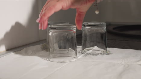 two clean glass cups placed upside down on white tissue to dry on countertop with third glass cup being carefully positioned under bright kitchen lighting