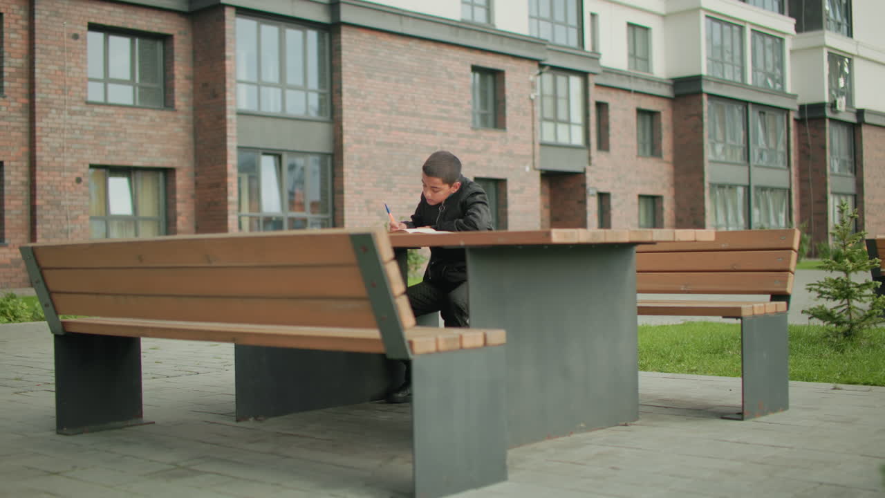 Young boy seated at outdoor bench near brick residential building focusing on writing in notebook with open book in front of him, creating scene of learning in urban environment