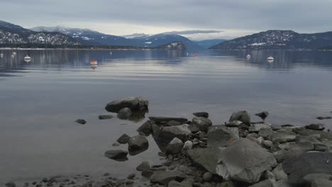 Shuswap-Lake-on-a-windless-day-with-low-hanging-clouds-with-Rocky-mountains-on-the-background-and-stones-on-the-foreground