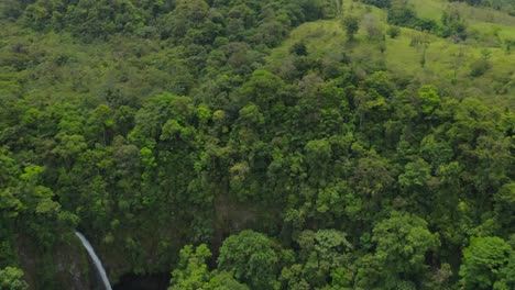 Fortuna-waterfall-in-lush-rainforest-valley-and-view-of-Arenal-volcano,-aerial