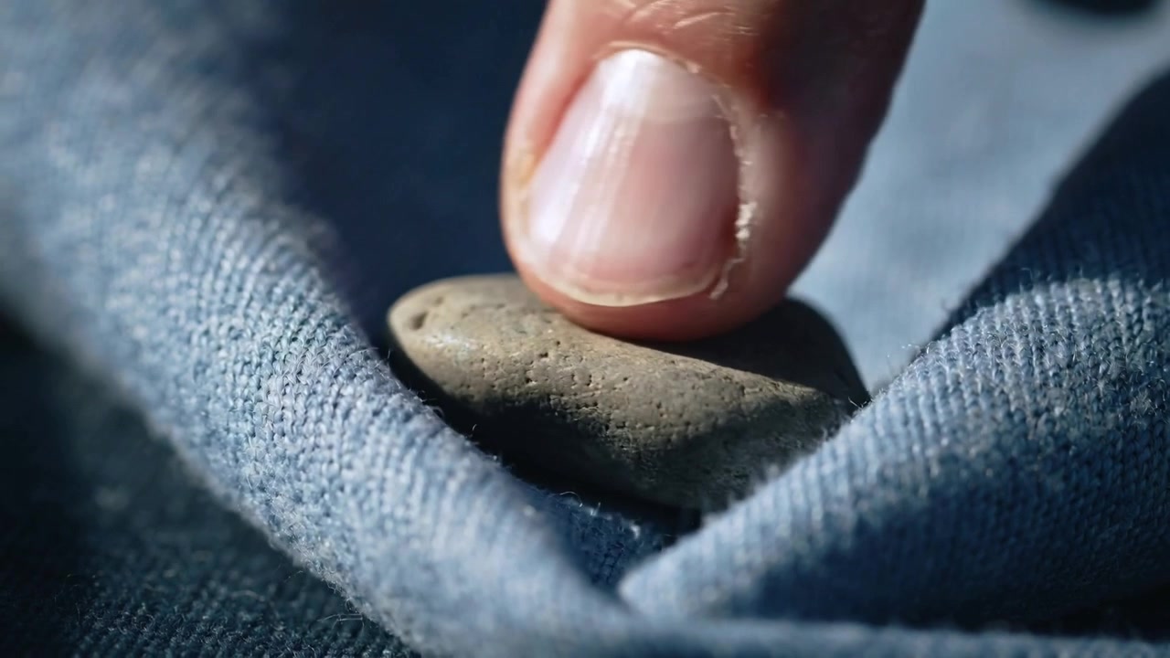 Approaching fingertip contacting and seating pebble into blue denim fold, showing pores