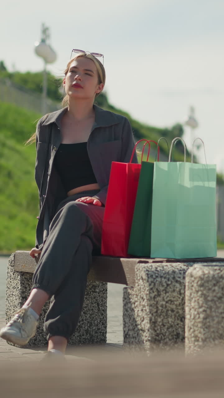 회색 옷을 입은 여자가 앉아있는 동안 벤치에 다채로운 쇼핑 가방을 떨어뜨립니다. 초록색 언덕과 배경에서 는 사람들은 약간 흐릿합니다.