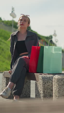 dama en ropa gris cae bolsas de compras de colores en el banco mientras se sienta, colinas de verdor y la gente caminando en el fondo son ligeramente borrosos