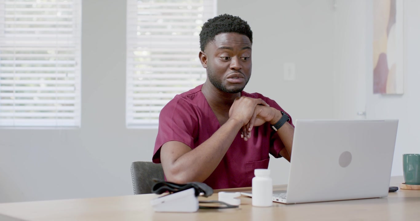African American male in burgundy scrubs sitting at table, speaking with laptop after call alert