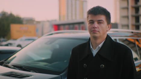 banker standing close to car with subtle smile, background shows parked cars and distant building at dusk, portraying confident business professional enjoying success in an urban setting