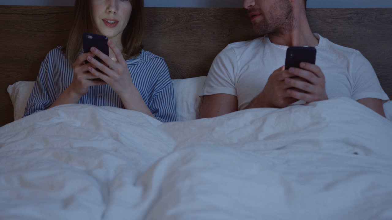 pareja caucásica usando teléfonos inteligentes mientras están sentados en la cama debajo de la manta