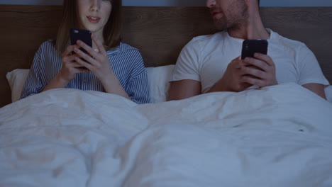 Caucasian-couple-using-smartphones-while-they-are-sitting-in-the-bed-under-the-blanket