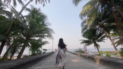 Slow-motion-shot-of-a-young-happy-woman-running-towards-the-ocean-surrounded-by-exotic-palm-trees,-rising-her-arms-in-the-sky-just-above-her-head-and-turnin-around-to-the-camera-smiling