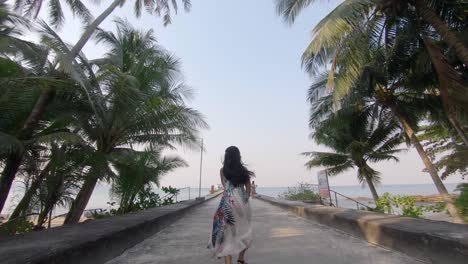 slow motion shot of a young happy woman running towards the ocean surrounded by exotic palm trees, rising her arms in the sky just above her head and turnin around to the camera smiling