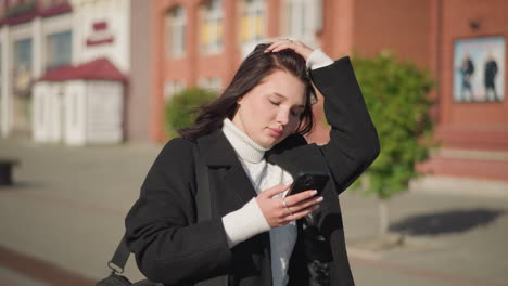 woman walking outdoors focused on her phone, adjusting hair back with sunlight reflecting off her face, background features red and white buildings and greenery under clear blue sky