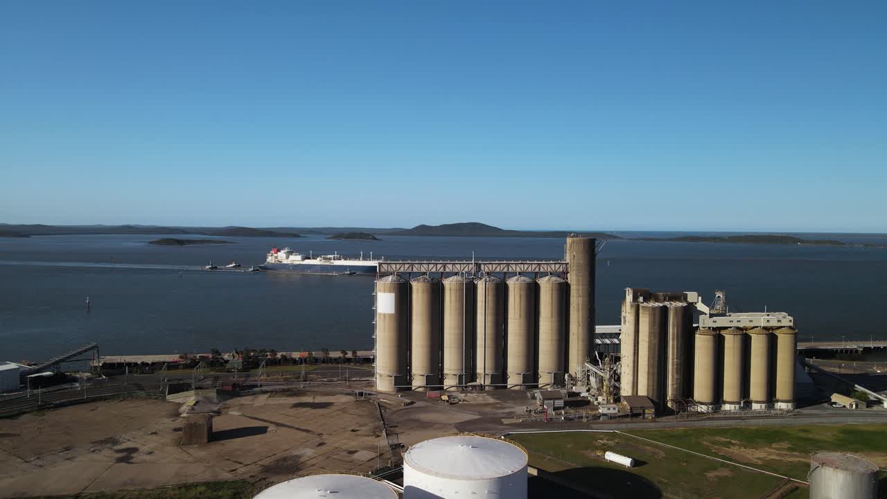 Um grande navio de carga navega pelas águas de um porto industrial, próximo a uma imponente instalação de armazenamento portuário. Imagem de drone em movimento.