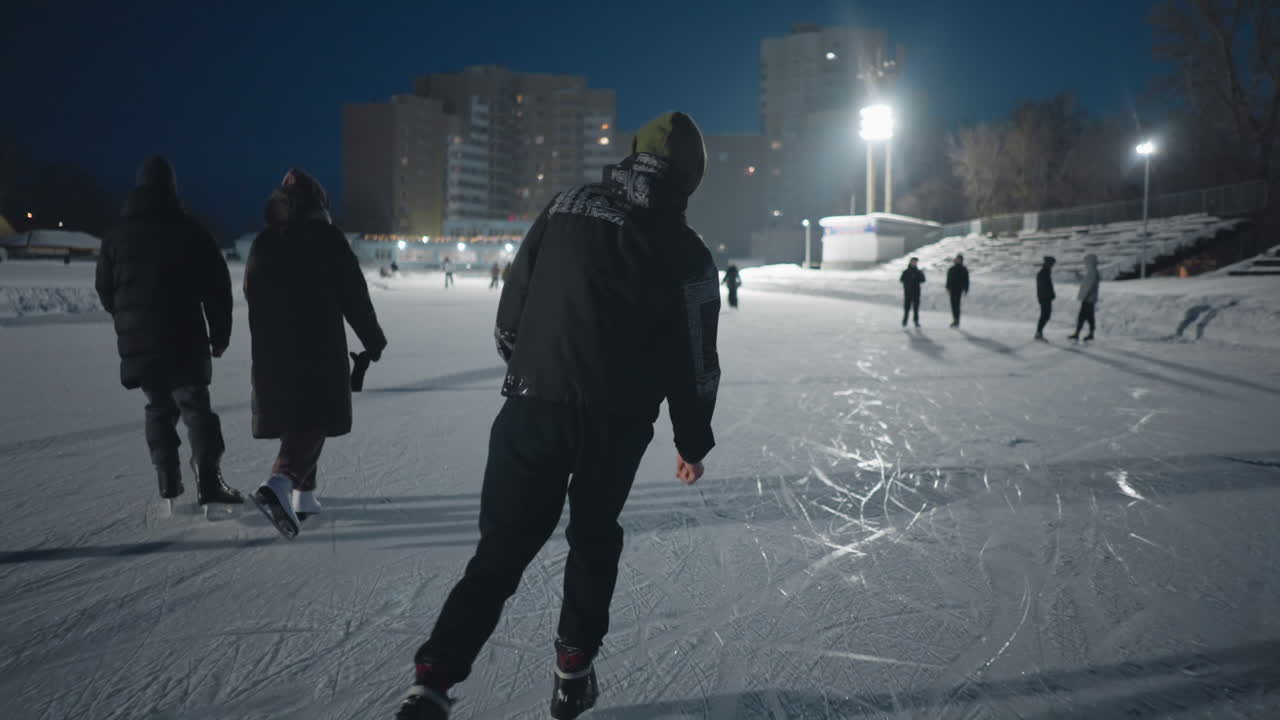 back view group of people skating together on outdoor ice rink at night under bright stadium lights visible urban buildings in background with skaters of all ages gliding across frozen surface