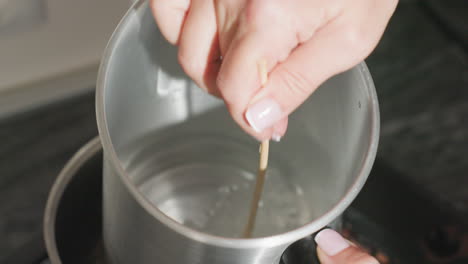 close up of hands stirring soluble substance in metal jug placed inside steaming pot on gas burner, heat creating gentle bubbles as solution dissolves in hot water during kitchen preparation process