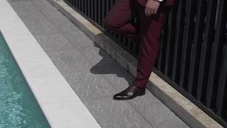 groom dressed in a stylish burgundy suit leans casually against a black fence by a poolside, exuding confidence and elegance