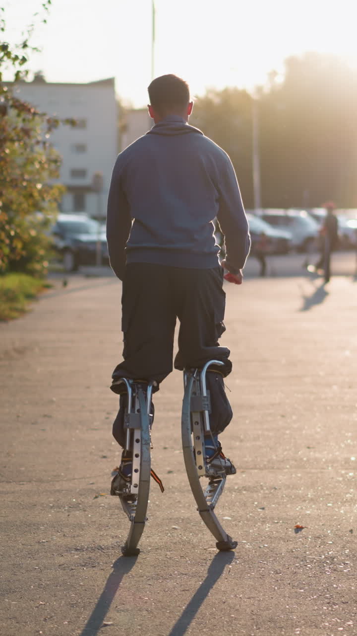 집과 사람들로 둘러싸인 주거 지역에서 해가 지는 동안 회색 후디와 어두운 회색 바지를 입고 손을 펴고 기둥에 뛰어드는 남자의 뒷면, 에너지, 균형을 보여줍니다.