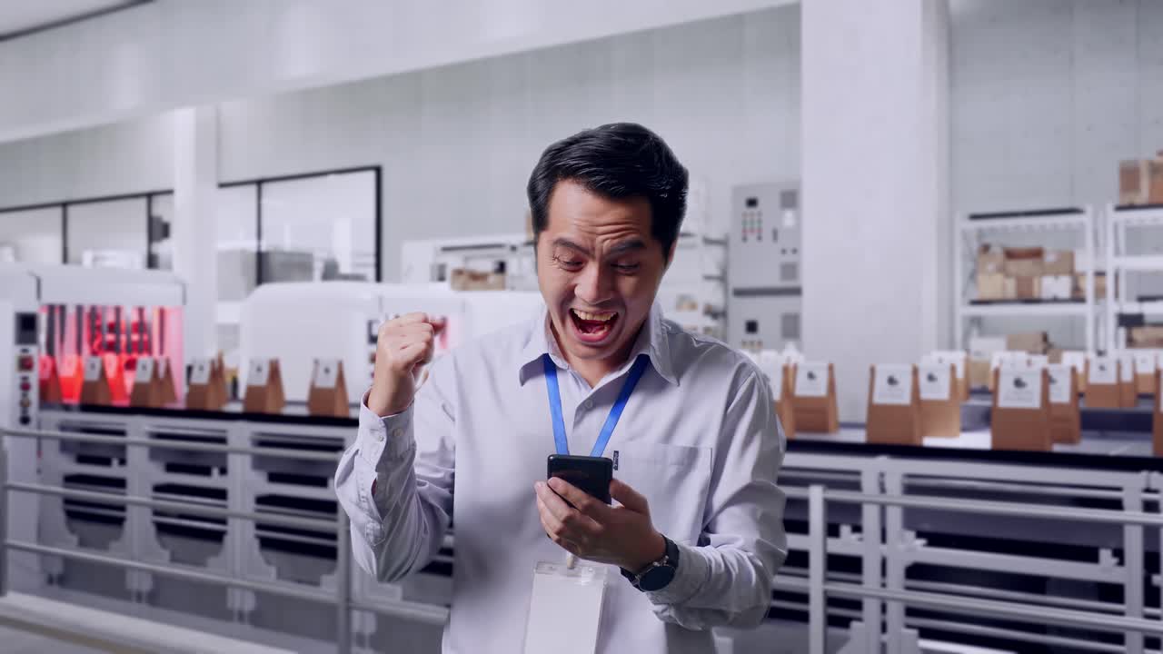 Asian Male Professional Worker Standing With His Smartphone With Conveyor for Packaging Coffee Beans in Bags at Coffee Factory, He Raises His Fist Up With Screaming Goal