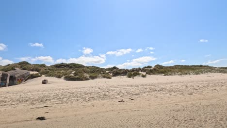 camera pans across sandy beach revealing historic, graffiti-covered world war ii bunkers under bright daylight, with clear blue sky and scattered clouds