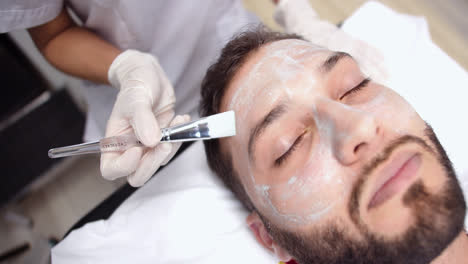 beautiful slow motion overhead shot of a young man lying on a massage table with his eyes closed in a dermatology clinic while having repair cream applied to him with a brush.