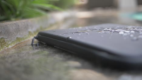 close up of a black, basic, waterproof laptop case with lines engraved in it lies on the side of a pool with splashes and drops of water on it