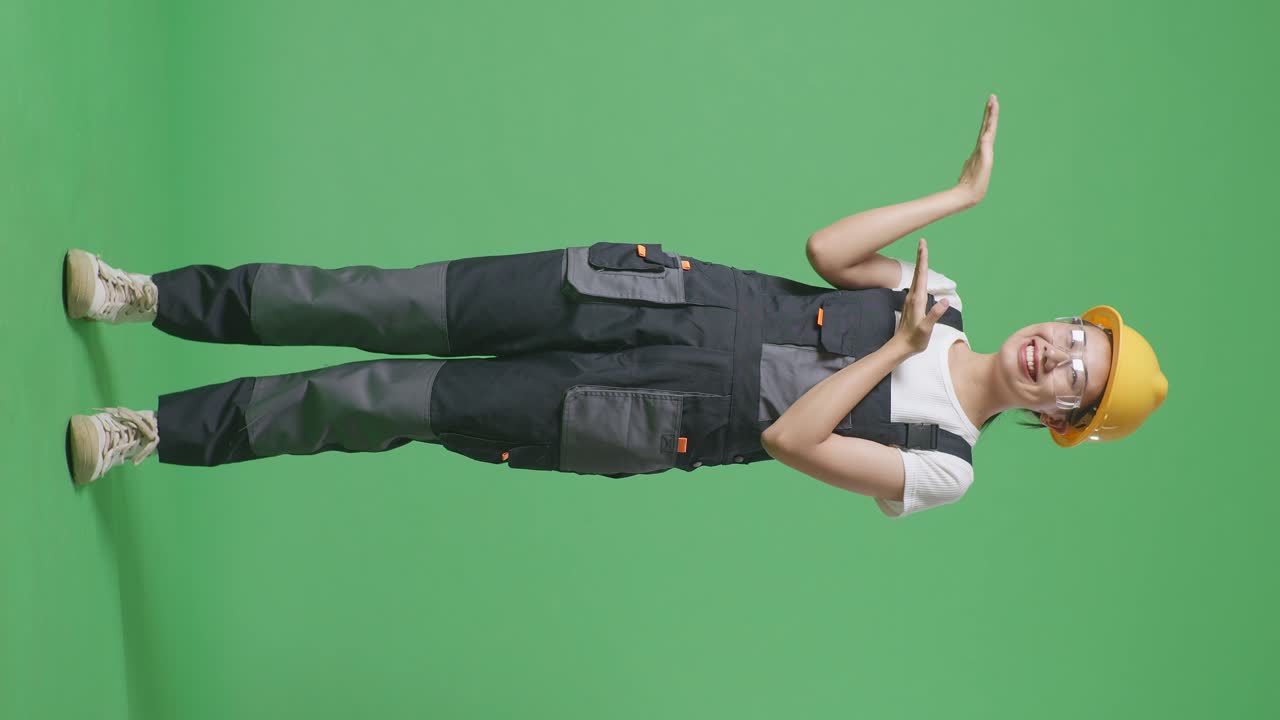 안경과 안전 헬을 입은 아시아 여성 노동자의 몸이 가득 차 있으며, 녹색 스크린 배경 스튜디오에 서서 웃고 을 가리키고 있습니다.