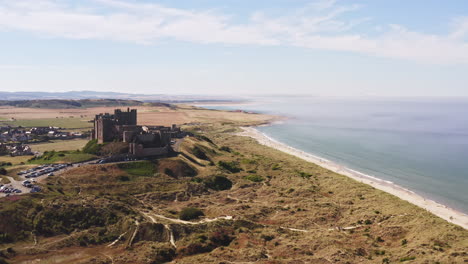 Aerial-shot-rising-over-sand-dunes-revealing-a-beautiful-castle-by-the-sea,-with-holidaymakers-on-the-beach-in-the-distance