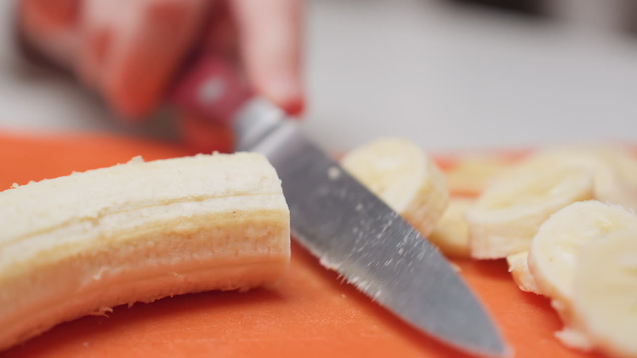 Close up hand cutting banana with knife on orange rubber board, round slices forming on counter, soft texture visible, shallow focus, kitchen prep mood, clean surface, fresh fruit ready for snack