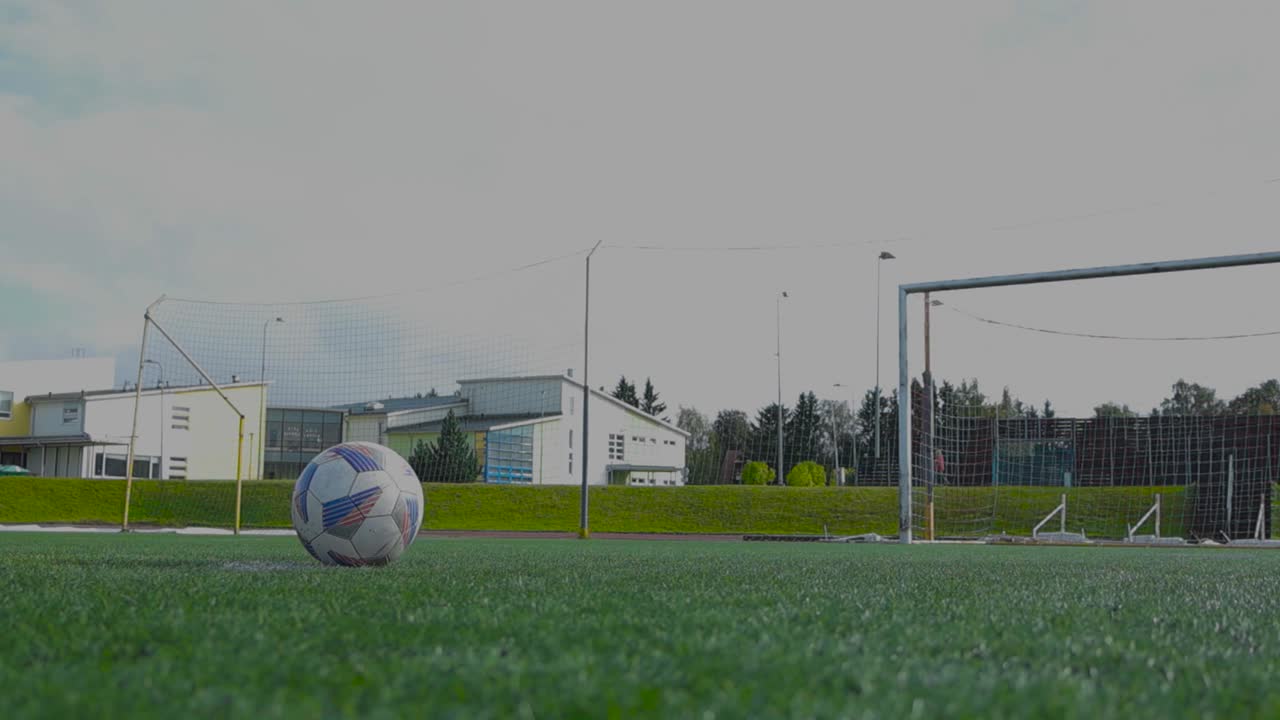 Person kicking a soccer ball or a football in a stadium making it go into the goal in front of him in slow motion. Video is filmed from the ground and Stadium short grass is visible with markings.