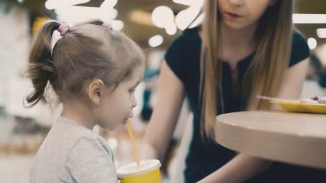 Young-beautiful-mother-with-her-little-daughter-sitting-in-a-cafe-Mom-gives-her-baby-drink-through-the-straw-teaches-a-child-to