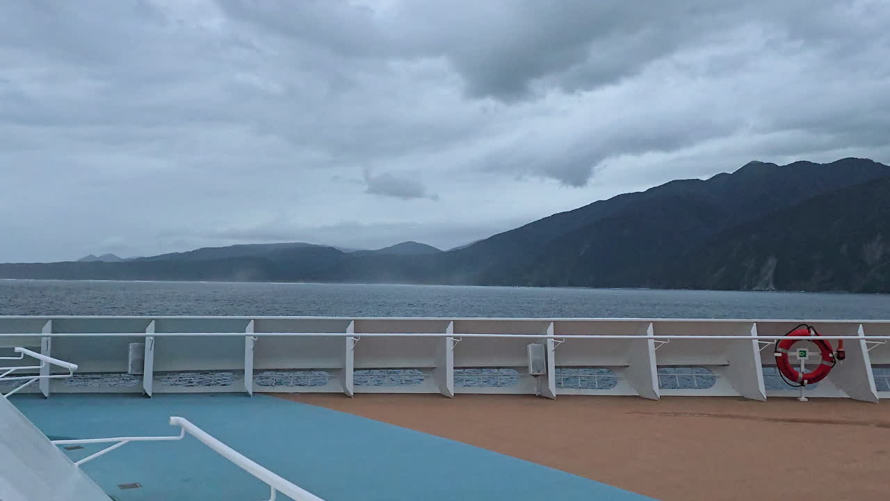 Cruise ship deck providing vantage point over Milford Sound, capturing the impressive fjord with towering mountains, gloomy sky, and vast body of water