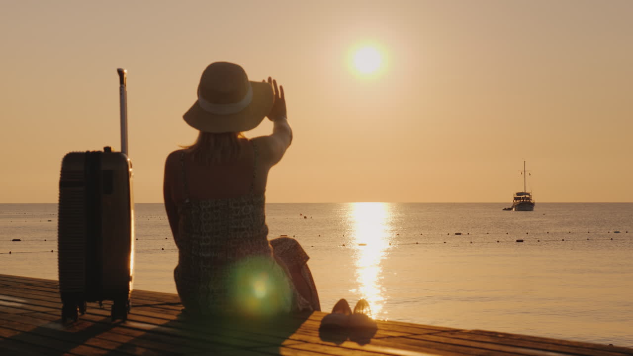 한 여성이 여행 개념인 그녀의 짐 근처 부두에 앉아 멀리 있는 배를 바라본다