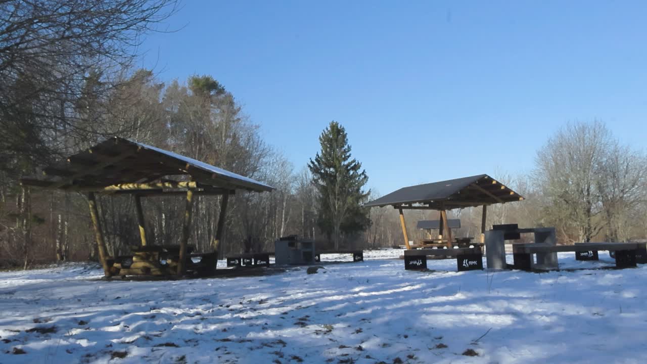 Calming scene of hiking trail vacation log and wooden picnic tables and grilling spot in winter nature during a gorgeous sunny day. The structures are modern and leafless bare trees surround them.