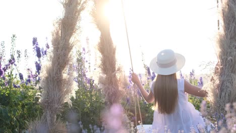 a young woman is riding on a swing at sunset she is wearing a beautiful dress and a hat a young woman is riding on a swing at sunset she is wearing a beautiful dress and a hat summer nature beautiful purple flowers back view