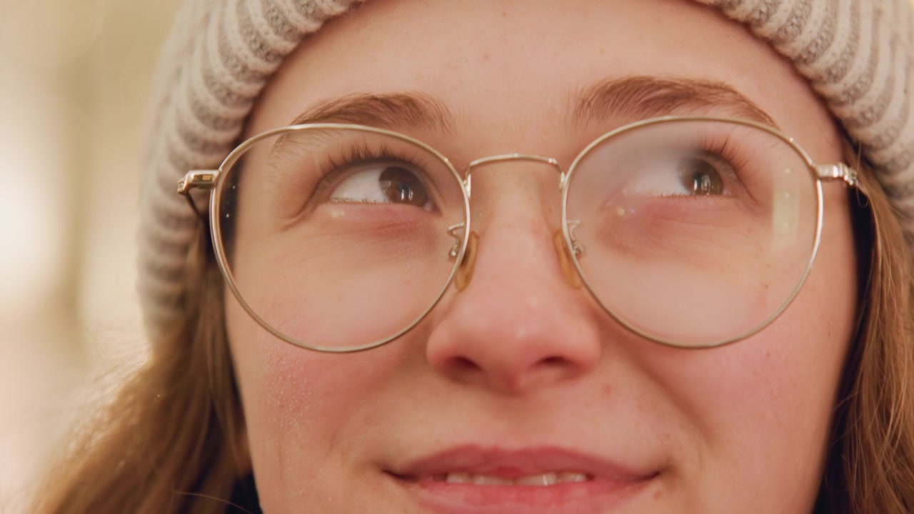 Woman with glasses smiling happily upward. Cheerful female in glasses gazes joyfully overhead. Female wearing spectacles beams in winter gear as she looks happily upward and smiles brightly