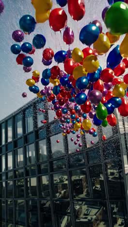 colorful balloons are flying away from a modern glass skyscraper in a generic urban city district with other buildings and trees visible in the background during a sunny day