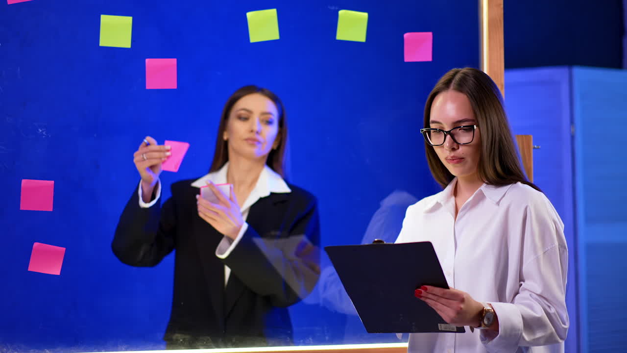 Long-haired girl with clipboard in hands stands at foreground. Woman wearing black jacket writes on sticker notes and puts them on glass wall.