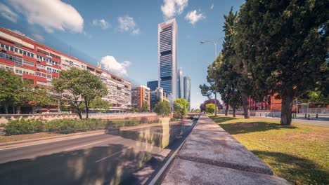 Toma-De-Tiempo-Ultra-Gran-Angular-De-Los-Rascacielos-De-Madrid-Contra-El-Cielo-Azul-Con-Nubes-Blancas-En-Una-Mañana-Soleada-De-Primavera-Y-Primer-Plano-De-Los-Autos-Que-Pasan