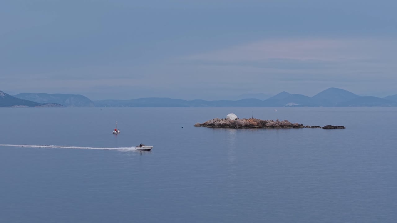 A speed boat glides across the sea, passing in front of the island chapel of St. Nikolaos near Hydra, Greece. Drone footage captures motion, tradition, and stunning scenery