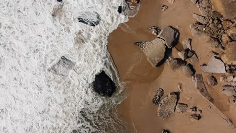 aerial top down shot of foamy waves reaching golden beach with rocks in portugal at sunny day